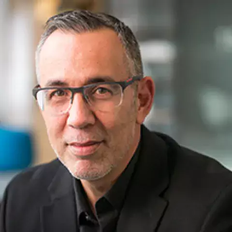 Headshot of A.J. Montero in black buttoned shirt and black framed glasses, against blurred background of office interior