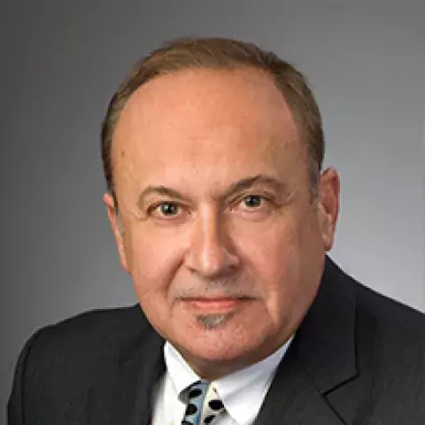 Headshot of Bob Falcone in black suit jacket with white shirt and patterned tie, against a gradated gray background