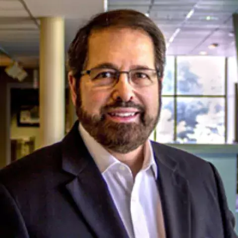 Headshot of Cameron James in a black suit jacket and white buttoned shirt, against the background of an office interior