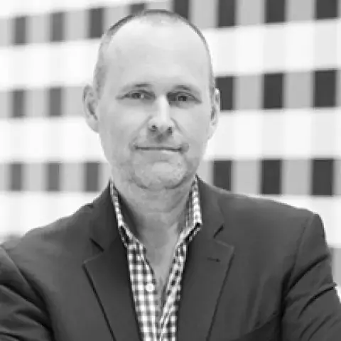 Black and white headshot of Tom Ramsey in dark jacket over a patterned buttoned shirt against a geometric patterned background